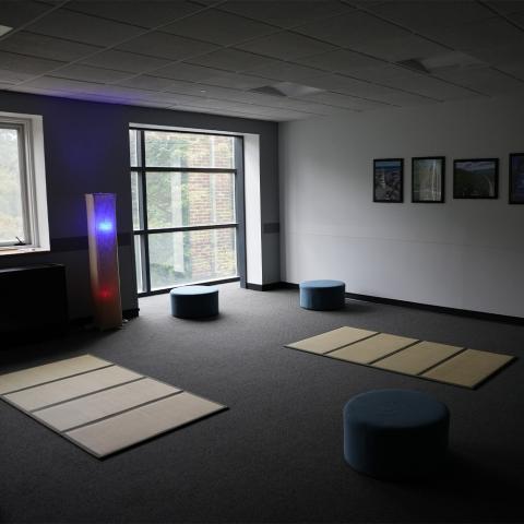 Room with mats on floor and round blue cusions, framed photos on wall, and a tall lamp with red and blue lights near large windows.